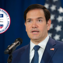 Marco Rubio habla frente audiencia frente a micrófono. Logo de USAID.