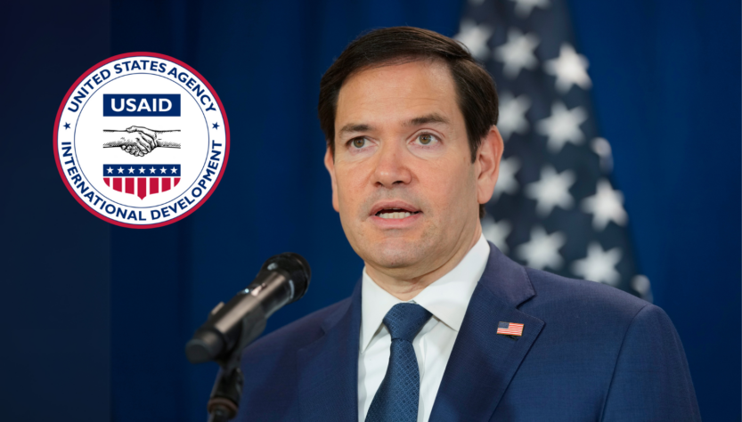 Marco Rubio habla frente audiencia frente a micrófono. Logo de USAID.