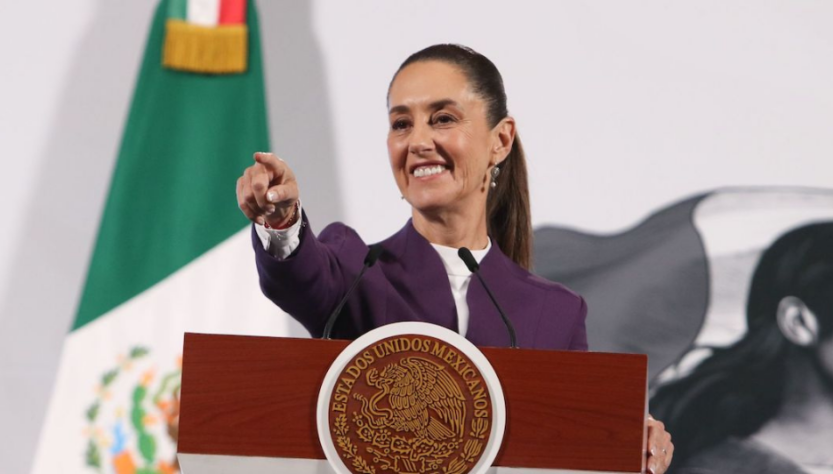 Claudia Sheinbaum frente a podio, sonríe y apunta con el dedo índice. conferencia